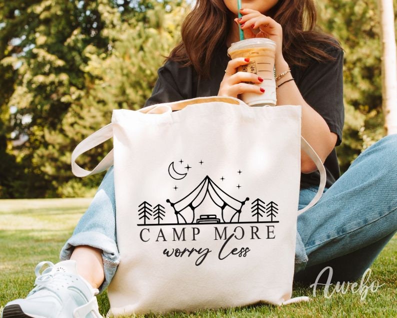 K&ouml;nnte beinhalten: Eine wei&szlig;e Canvas-Tragetasche mit schwarzem Text, der "Camp More Worry Less" lautet. Die Tasche zeigt eine Schwarzwei&szlig;illustration eines Zeltes, Sterne und B&auml;ume.