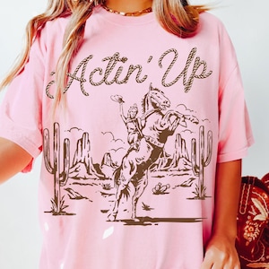 May include: Pink t-shirt with a brown graphic of a woman riding a bucking horse, cacti, and desert landscape. The text "Actin' Up" is above the graphic in a rope-like font.