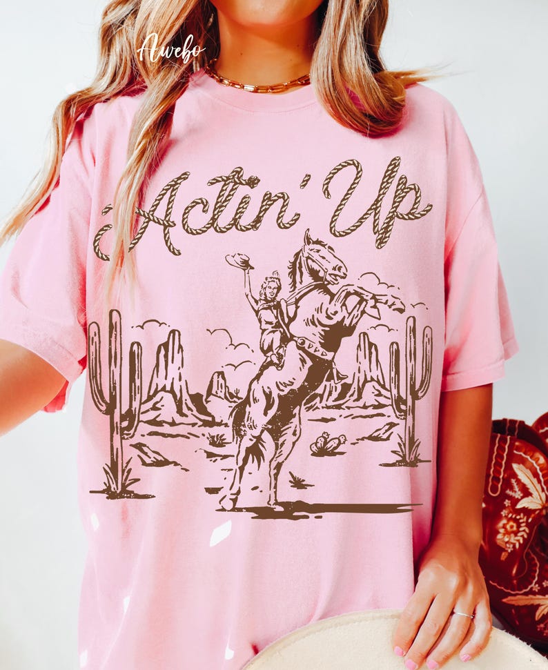 May include: Pink t-shirt with a brown graphic of a woman riding a bucking horse, cacti, and desert landscape. The text "Actin' Up" is above the graphic in a rope-like font.