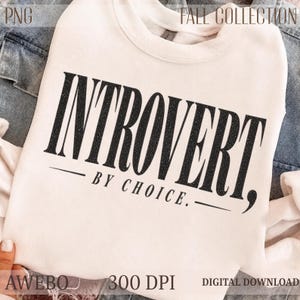 May include: A cream-colored crewneck sweatshirt featuring the black text "INTROVERT, BY CHOICE." printed in a bold, serif font. The design includes horizontal lines above and below the smaller text. The sweatshirt is displayed against a denim background.