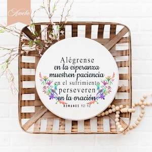 Puede incluir: Un letrero redondo blanco con un dise&ntilde;o floral y el texto "Al&eacute;grense en la esperanza muestren paciencia en el sufrimiento perseveren en la oraci&oacute;n ROMANOS 12:12". El letrero est&aacute; colgado en una caja de madera.