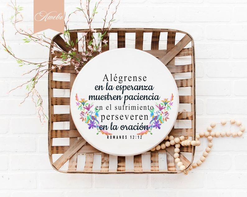 Puede incluir: Un letrero redondo blanco con un dise&ntilde;o floral y el texto "Al&eacute;grense en la esperanza muestren paciencia en el sufrimiento perseveren en la oraci&oacute;n ROMANOS 12:12". El letrero est&aacute; colgado en una caja de madera.
