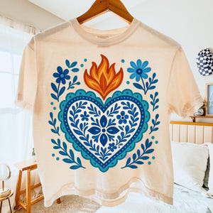 May include: A light peach-colored t-shirt with a heart-shaped design. The heart is outlined in blue with a dotted border and filled with blue floral patterns. Above the heart is a flame design in orange and yellow. Blue floral accents surround the heart.
