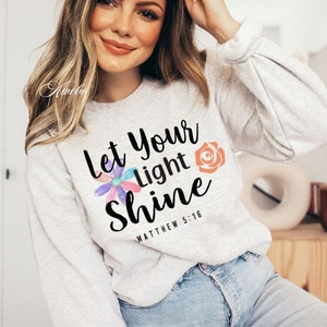 May include: A gray sweatshirt with the text "Let your light shine" and the scripture reference "Matthew 5:16". The text is in black and is surrounded by colorful flowers.