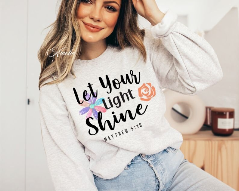 May include: A gray sweatshirt with the text "Let your light shine" and the scripture reference "Matthew 5:16". The text is in black and is surrounded by colorful flowers.