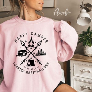 K&ouml;nnte beinhalten: Rosafarbener Pullover mit einem grafischen Design eines Wohnmobils, eines Lagerfeuers und B&auml;umen. Der Text lautet "Happy Camper Happiness is Toasted Marshmallows".