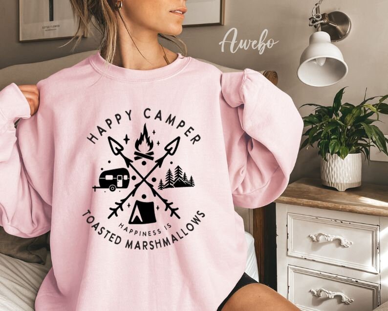K&ouml;nnte beinhalten: Rosafarbener Pullover mit einem grafischen Design eines Wohnmobils, eines Lagerfeuers und B&auml;umen. Der Text lautet "Happy Camper Happiness is Toasted Marshmallows".
