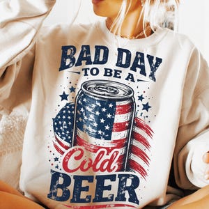 May include: Cream-colored sweatshirt with a graphic design featuring a beer can decorated with the American flag. The text reads "BAD DAY TO BE A COLD BEER" in navy blue and red.