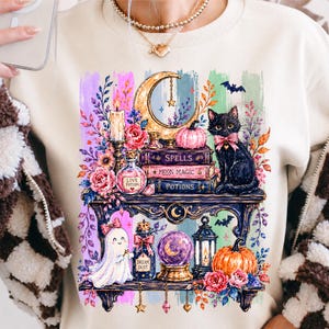 May include: A beige sweatshirt featuring a graphic print of two shelves filled with magical items including a black cat with a pink bow, a ghost, a crescent moon, pumpkins, books labeled Spells, Moon Magic, and Potions, and glass bottles.