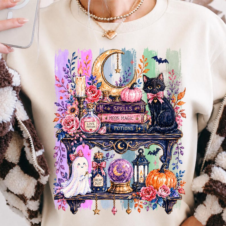 May include: A beige sweatshirt featuring a graphic print of two shelves filled with magical items including a black cat with a pink bow, a ghost, a crescent moon, pumpkins, books labeled Spells, Moon Magic, and Potions, and glass bottles.