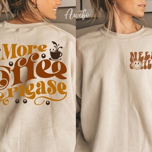 Może przedstawiać: Dwie beżowe bluzy z grafiką związaną z kawą. Jedna bluza ma napis "More Coffee Please" z grafiką kubka z kawą. Druga bluza ma napis "Need Coffee" z grafiką uśmiechniętej twarzy.