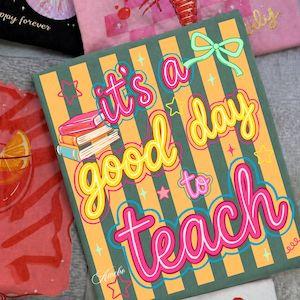 May include: A square, green and orange striped canvas with the text "It's a good day to teach" in bright colors. The design includes a stack of books, stars, and a bow. The artist's signature "Amelio" is at the bottom.