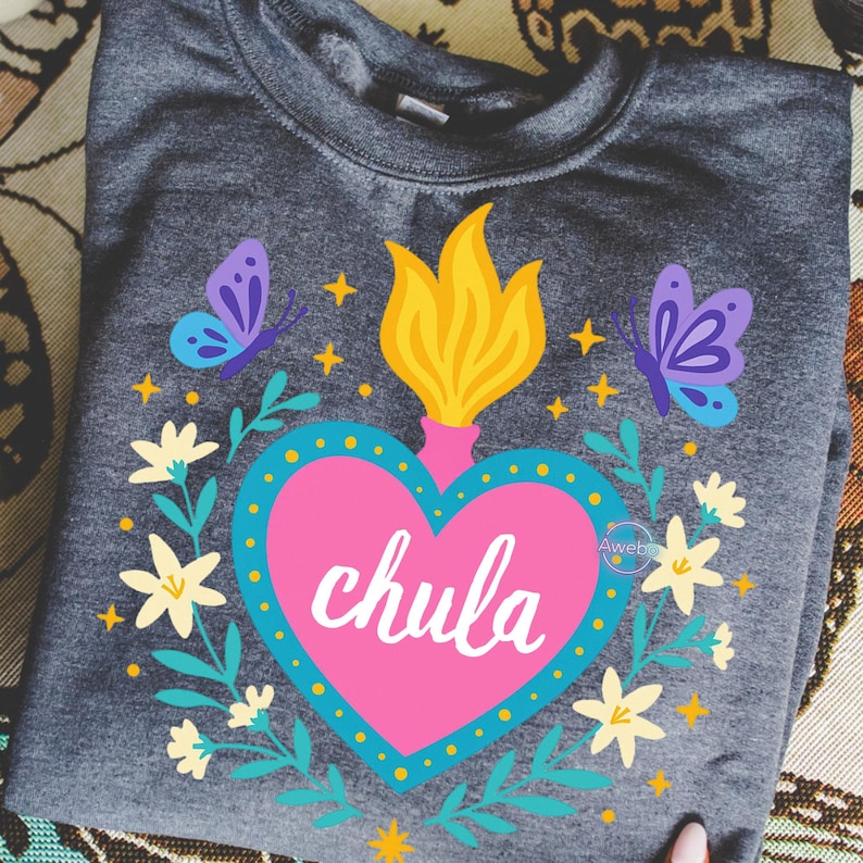 May include: A gray sweatshirt featuring a pink heart with the word "chula" in white script. The heart is surrounded by a blue dotted border, yellow flames, butterflies, and floral accents. The design is centered on the folded garment.