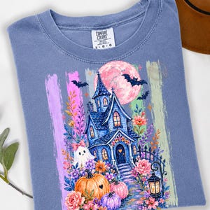 May include: A blue t-shirt featuring a graphic print of a haunted house, a pink moon, bats, pumpkins, and a ghost. The design includes colorful floral accents and a lantern. The shirt has a Comfort Colors brand label at the neck indicating size large.