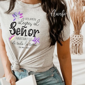 Puede incluir: Una camiseta gris con un gr&aacute;fico negro que dice "Aclamen alegres al Se&ntilde;or Habitantes de toda la tierra Salmo 100:1" con flores moradas y amarillas que rodean el texto.
