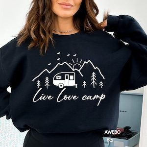 K&ouml;nnte beinhalten: Schwarzer Pullover mit einem wei&szlig;en Grafikdesign eines Wohnwagens, Bergen, einer Sonne und B&auml;umen. Der Text "Live Love Camp" steht unter der Grafik.