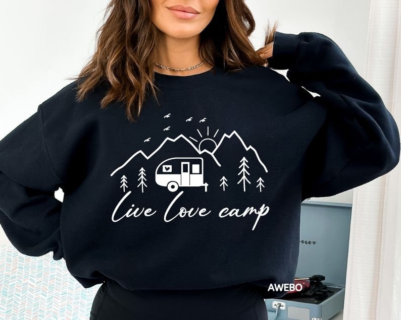 K&ouml;nnte beinhalten: Schwarzer Pullover mit einem wei&szlig;en Grafikdesign eines Wohnwagens, Bergen, einer Sonne und B&auml;umen. Der Text "Live Love Camp" steht unter der Grafik.
