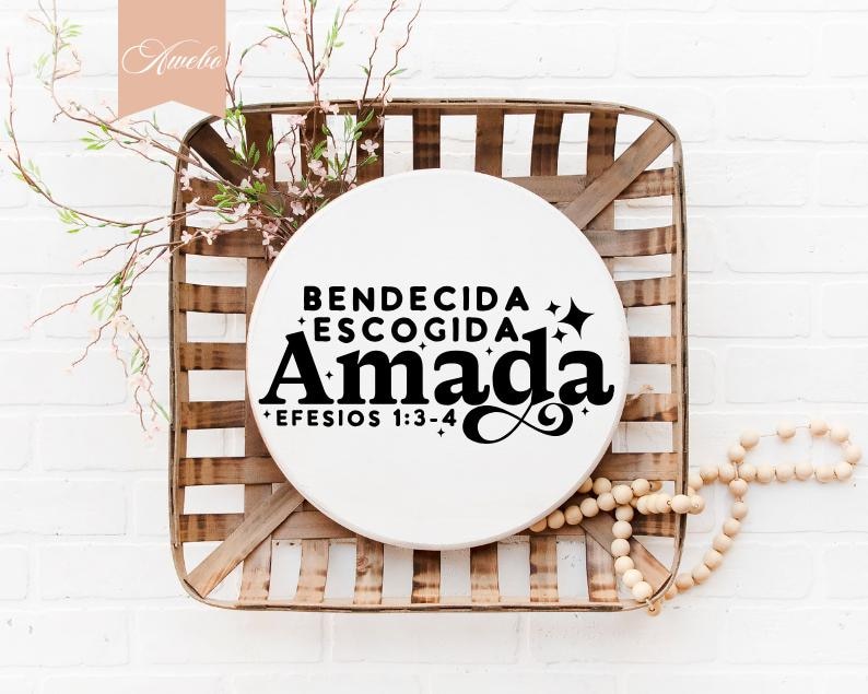 May include: A white, round sign with the words "BENDECIDA ESCOGIDA AMADA EFESIOS 1:3-4" in black lettering, set against a woven basket. The basket is brown and white, with floral accents and a string of wooden beads.