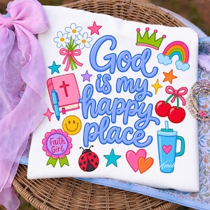 Pode incluir: Camiseta branca com a frase "God is my happy place" em azul. O design inclui um arco-íris, uma coroa, cerejas, uma bíblia, estrelas, uma joaninha e um smiley. As palavras "Faith Girl" também estão incluídas.