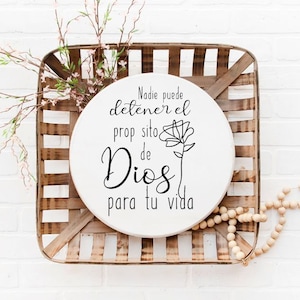 May include: A white round wooden sign with a black floral design and the text "Nadie puede detener el proposito de Dios para tu vida". The sign is sitting in a wooden basket.