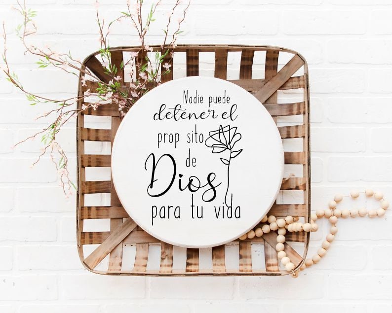 May include: A white round wooden sign with a black floral design and the text "Nadie puede detener el proposito de Dios para tu vida". The sign is sitting in a wooden basket.