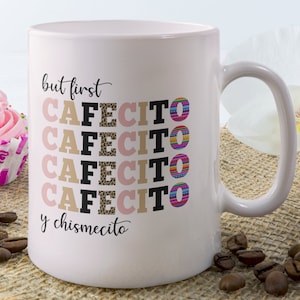 Puede incluir: Una taza de caf&eacute; de cer&aacute;mica rosa con el texto "but first cafecito y chismecito" en negro. La palabra "cafecito" se repite cuatro veces, cada una con un patr&oacute;n colorido diferente.