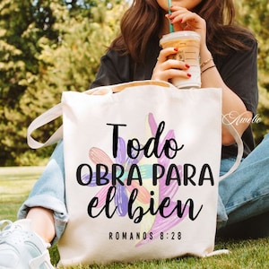 Puede incluir: Una bolsa de lona blanca con el texto "Todo obra para el bien" y "Romanos 8:28" en letras negras. La bolsa tiene un dise&ntilde;o floral con acentos rosas, morados y amarillos.