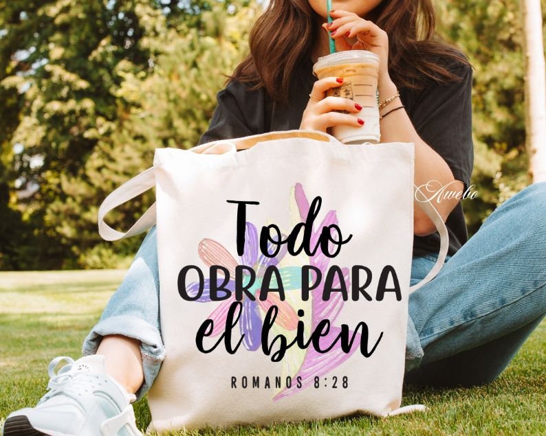 Puede incluir: Una bolsa de lona blanca con el texto "Todo obra para el bien" y "Romanos 8:28" en letras negras. La bolsa tiene un dise&ntilde;o floral con acentos rosas, morados y amarillos.