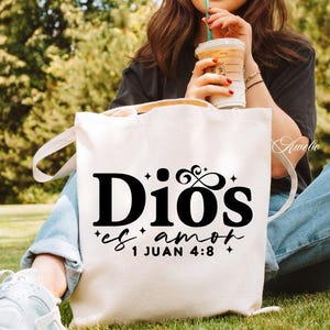 May include: A white canvas tote bag with black text that reads "Dios es amor 1 Juan 4:8". The bag has two handles and is being held by a person sitting outside. The person is wearing blue jeans and white sneakers.
