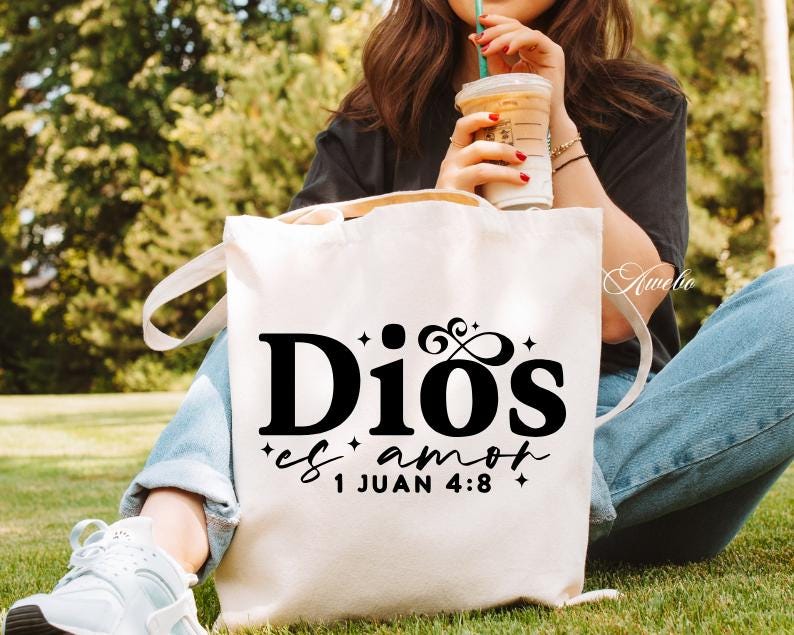 May include: A white canvas tote bag with black text that reads "Dios es amor 1 Juan 4:8". The bag has two handles and is being held by a person sitting outside. The person is wearing blue jeans and white sneakers.