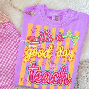 May include: A lilac Comfort Colors t-shirt with the phrase "it's a good day to teach" in a colorful, playful font. The design includes books, stars, and a bow. The shirt is paired with pink gingham shorts and a gold necklace.