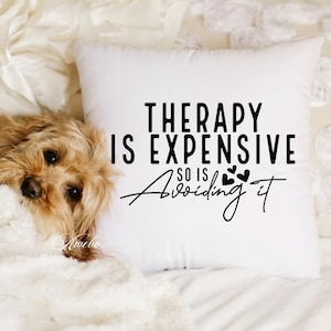 Puede incluir: Almohada blanca con texto negro que dice "La terapia es cara, tambi&eacute;n lo es evitarla". El texto est&aacute; rodeado de peque&ntilde;os corazones negros. Un perro marr&oacute;n y blanco est&aacute; acostado sobre una manta blanca en primer plano.
