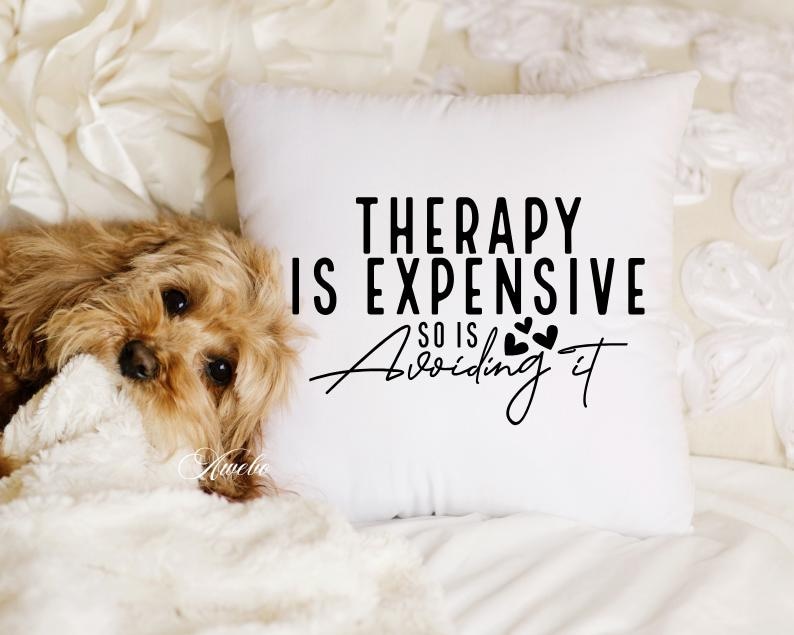 Puede incluir: Almohada blanca con texto negro que dice "La terapia es cara, tambi&eacute;n lo es evitarla". El texto est&aacute; rodeado de peque&ntilde;os corazones negros. Un perro marr&oacute;n y blanco est&aacute; acostado sobre una manta blanca en primer plano.