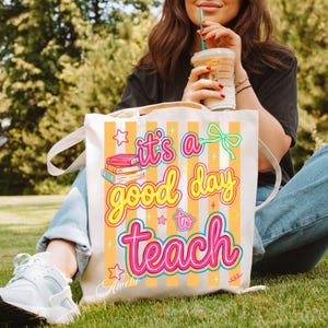 May include: Cream-colored tote bag with the text "it's a good day to teach" in pink and yellow. The bag has orange vertical stripes and a graphic of books. The bag has white handles.