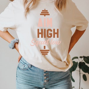 Pode incluir: Uma camiseta branca com um gr&aacute;fico marrom e rosa que diz "Aim High Squat Low" com um gr&aacute;fico de barra de halteres acima e abaixo do texto.
