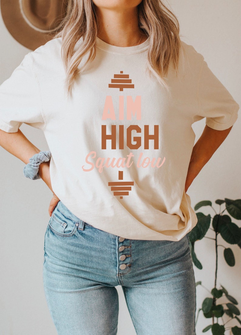 Pode incluir: Uma camiseta branca com um gr&aacute;fico marrom e rosa que diz "Aim High Squat Low" com um gr&aacute;fico de barra de halteres acima e abaixo do texto.