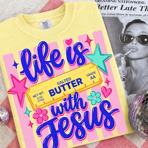 May include: Yellow t-shirt with the phrase "life is with Jesus" in blue script. The design includes a butter stick graphic, pink stars, hearts, and flowers. The shirt also has the text "Salted Butter" and "Net Wt. 4 oz. (113g)".