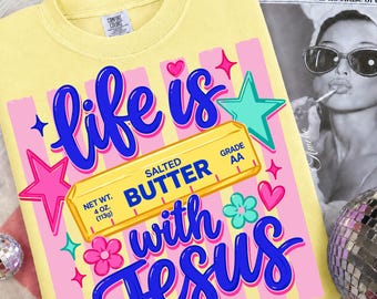 May include: Yellow t-shirt with the phrase "life is with Jesus" in blue script. The design includes a butter stick graphic, pink stars, hearts, and flowers. The shirt also has the text "Salted Butter" and "Net Wt. 4 oz. (113g)".