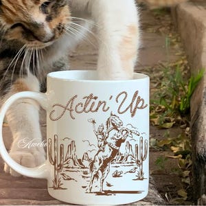 May include: White ceramic mug with the text "Actin' Up" in a brown script font. The mug features a brown illustration of a cowboy riding a bucking bronco, with cacti and a desert landscape in the background.