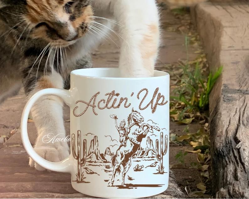 May include: White ceramic mug with the text "Actin' Up" in a brown script font. The mug features a brown illustration of a cowboy riding a bucking bronco, with cacti and a desert landscape in the background.
