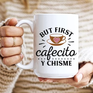 May include: White ceramic mug with black text that reads "But first cafecito y chisme". The mug has a brown illustration of a coffee cup with a pink heart-shaped pastry inside.