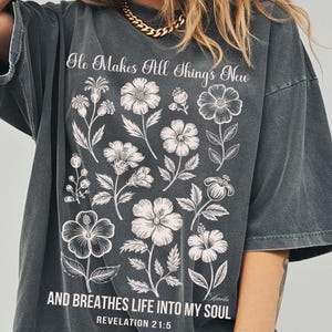 May include: Dark gray t-shirt with a floral design and the text "He Makes All Things New" and "AND BREATHES LIFE INTO MY SOUL" from Revelation 21:5. The shirt features a crew neck and short sleeves.