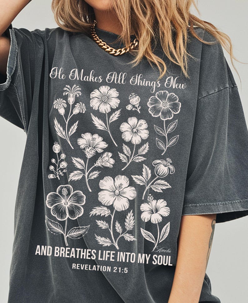 May include: Dark gray t-shirt with a floral design and the text "He Makes All Things New" and "AND BREATHES LIFE INTO MY SOUL" from Revelation 21:5. The shirt features a crew neck and short sleeves.