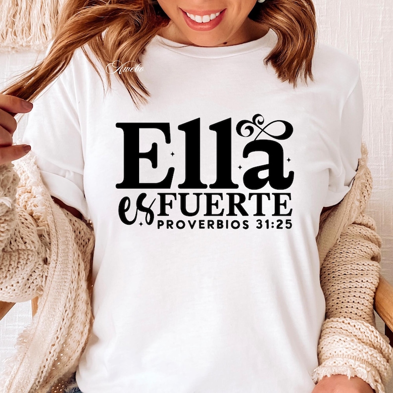 May include: White t-shirt with the text "Ella es FUERTE PROVERBIOS 31:25" in black. The shirt has short sleeves and a crew neck. The text is in a bold, stylized font.