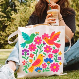 May include: A cream-colored tote bag featuring a colorful floral and bird design. The design includes three birds in green, orange, and yellow, surrounded by pink, purple, and blue flowers. The bag has two white handles.