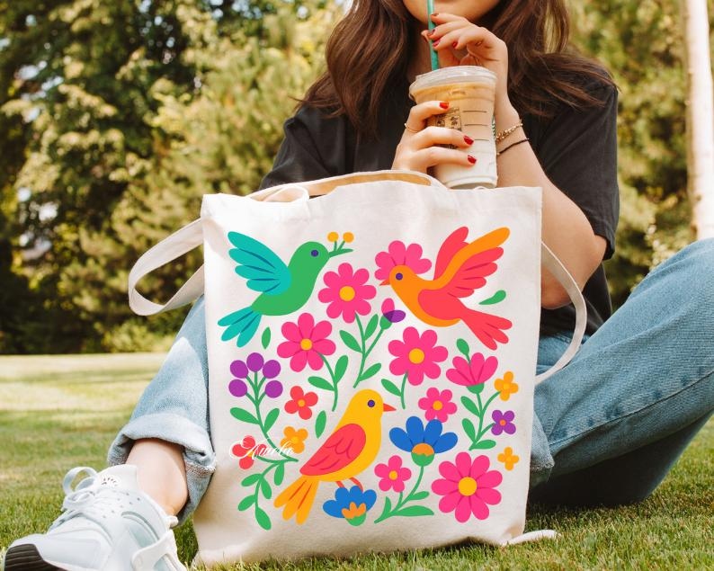 May include: A cream-colored tote bag featuring a colorful floral and bird design. The design includes three birds in green, orange, and yellow, surrounded by pink, purple, and blue flowers. The bag has two white handles.