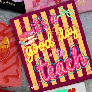 May include: A burgundy notebook with the text "it's a good day to teach" in a colorful, playful font. The design includes books, stars, and a bow, set against a striped orange and burgundy background. The notebook is likely for teachers.