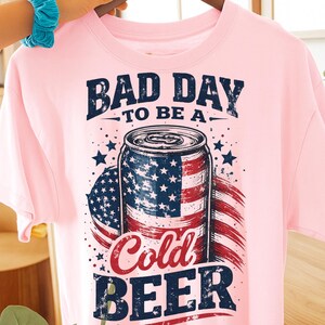 May include: A light pink t-shirt featuring the text "BAD DAY TO BE A..." above a graphic of a beer can with an American flag design. The words "Cold BEER" are printed below the can. The shirt is hanging on a wooden hanger.