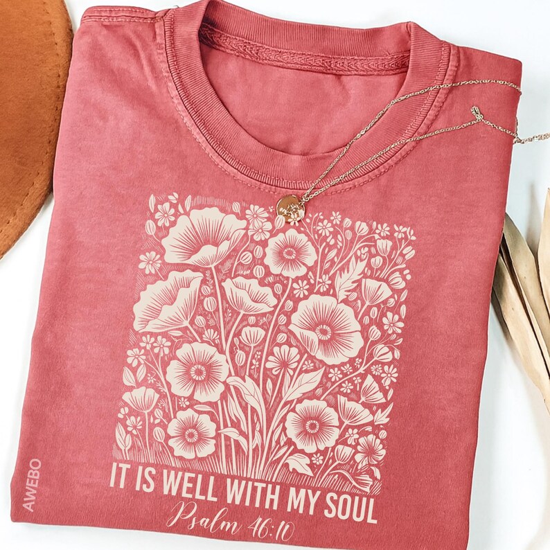 May include: A red-orange t-shirt with a floral design and the text "IT IS WELL WITH MY SOUL Psalm 46:10". A gold necklace with a pendant rests on the shirt. The shirt has a crew neck and short sleeves.