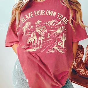 May include: A maroon t-shirt with the text "BLAZE YOUR OWN TRAIL" above a graphic of a person riding a horse through a desert landscape. The design includes mountains, cacti, and a winding road.